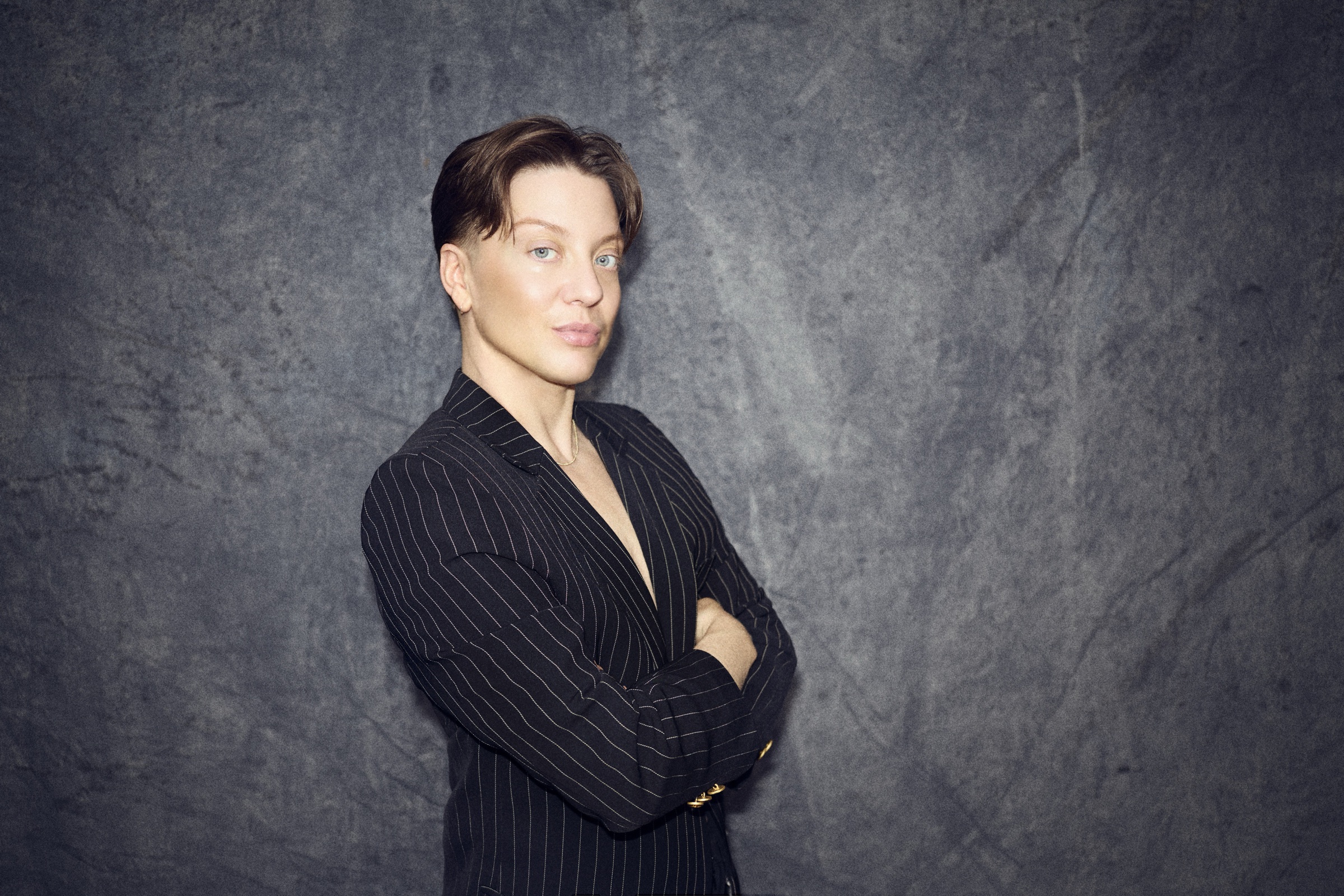K. Iver in a pinstripe jacket, arms crossed, against grey